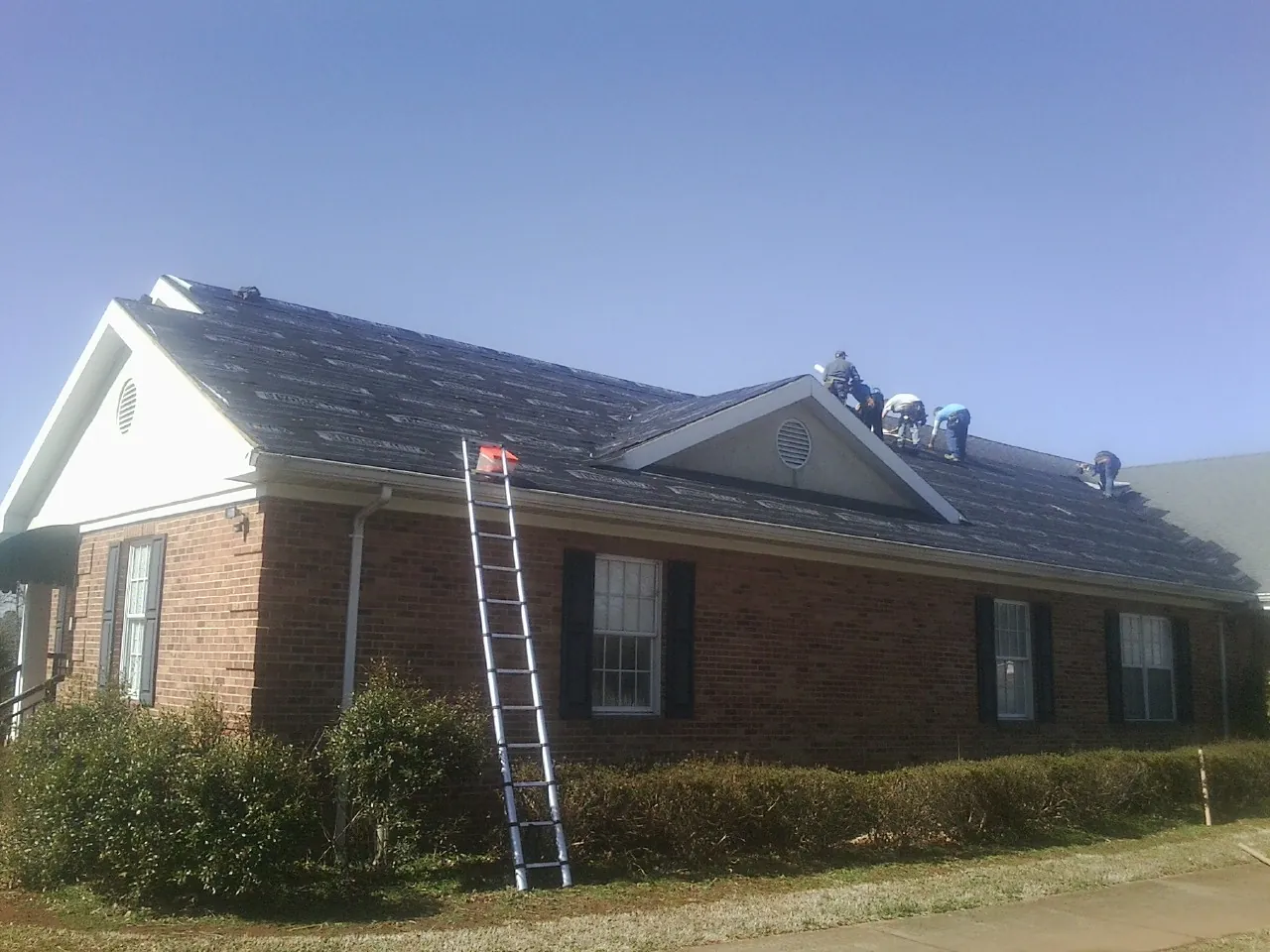 Crew tearing shingles off a brick ranch roof with a ladder set — Emergency Roof Repair work by our crew in Lehman