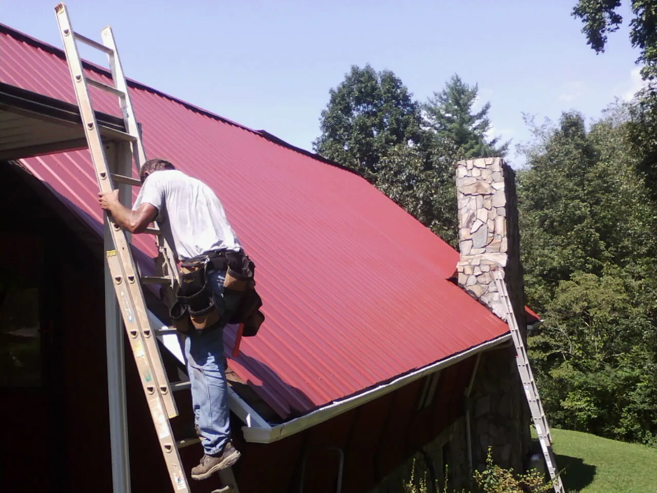 Roofer on a ladder beside a red metal roof and stone chimney — Soffit & Fascia Repair by Roofing Brothers in Lehman